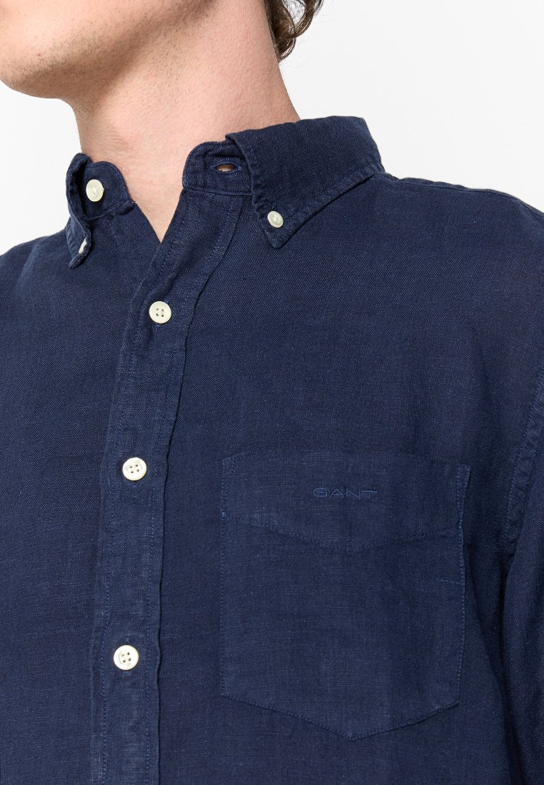 Camisa azul marino de botones hecha de lino, con un bolsillo, cuello redondeado y botones cremosos en contraste. Presenta un sutil bordado del logo de la marca.