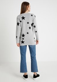 Sweater cinza de mangas longas com padrões de estrelas pretas nas costas, combinado com jeans azuis de boca de sino e sapatilhas pretas.