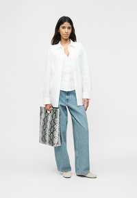 Γυναίκα με λευκό πουκάμισο και μπλε τζιν κρατώντας μια τσάντα ώμου με μοτίβο δέρματος φιδιού σε μαύρο και άσπρο, στέκεται μπροστά σε απλό φόντο.