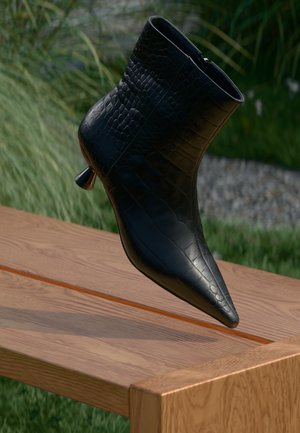 Stivale ankle in pelle nera con texture coccodrillo, punta affusolata e un accento metallico su un tacco sottile, posato su una superficie di legno.