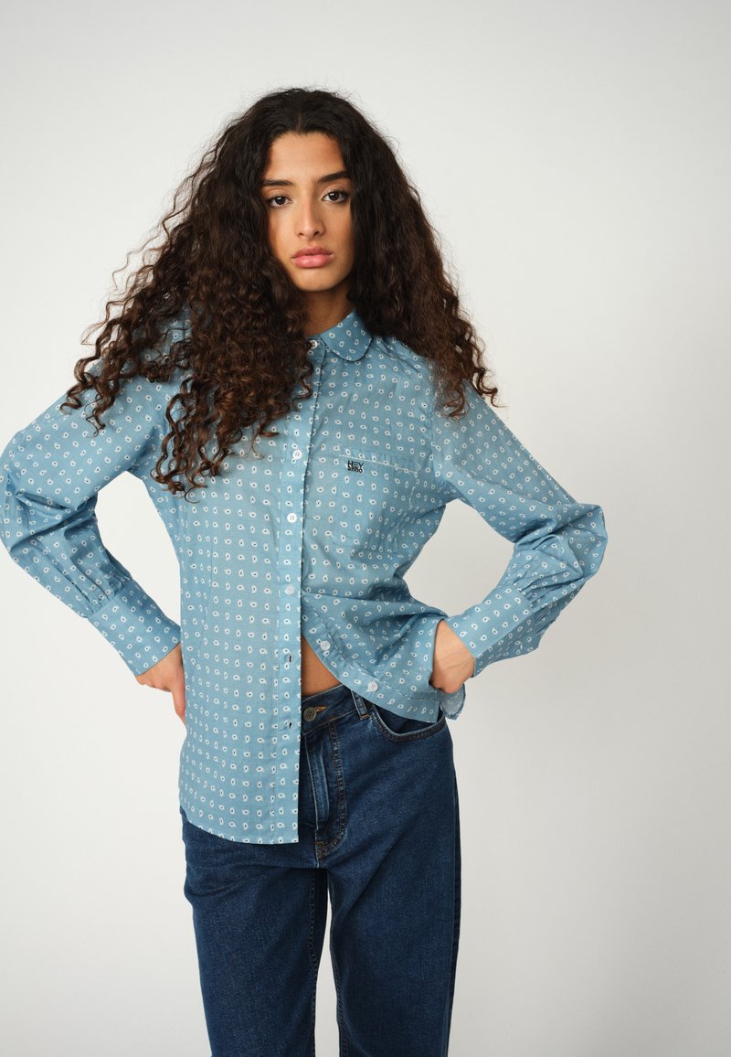 Light blue button-up shirt with a collar, featuring a small white pattern. Paired with dark blue jeans, showing a relaxed fit.