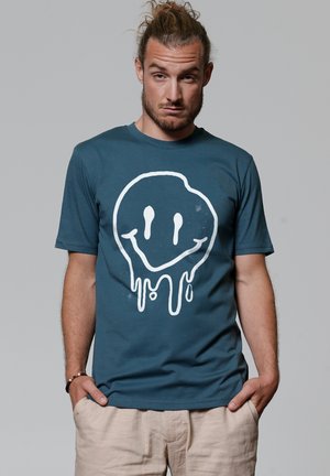 Teal katoenen T-shirt met een grote witte smiley-gezichtgrafiek met druipende details. Korte mouwen en een ronde hals.