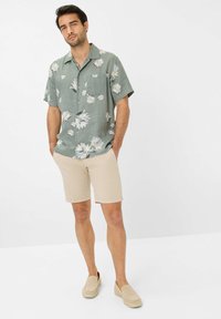 Camicia a maniche corte di colore verde chiaro con un motivo floreale, abbinata a pantaloni corti beige e scarpe slip-on beige. Tessuto con texture e dettaglio della tasca.