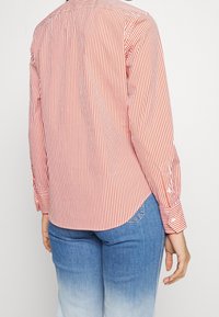 Red and white striped button-up shirt with long sleeves, a rounded hem, and button cuffs, made of a lightweight woven material.