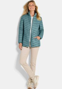 Woman standing with one hand in the pocket of a teal quilted jacket, wearing beige pants and beige sneakers, against a plain background.