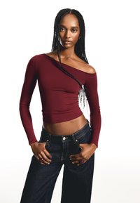 Fiatal nő hosszú fonatokkal, bordó off-shoulder crop toppot és sötét, magas derekú farmert viselve áll, keze a zsebében.