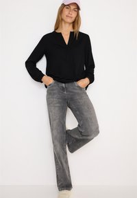 Schwarzes V-Ausschnitt Langarmshirt mit elastischen Bündchen, kombiniert mit verwaschenen grauen Schlagjeans und beigen Sneakers, akzentuiert durch eine hellrosa Mütze.