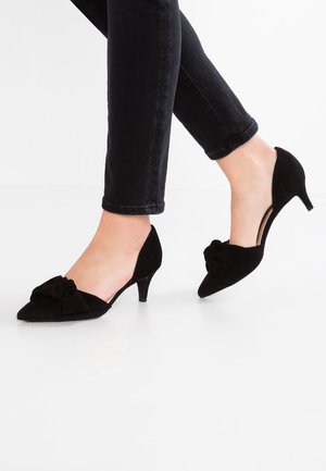 Escarpins en daim noir à petit talon, bouts pointus et détail nœud, portés avec un pantalon noir ajusté sur un sol blanc propre.