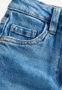 Jean en denim avec un délavage bleu clair, doté d'une poche arrière standard, de rivets en cuivre et de surpiqûres contrastantes marron à la taille et sur les coutures.