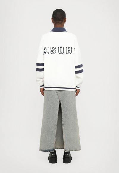 White sweatshirt with "KSUBI" lettering on the back, navy collar and striped accents. Paired with a long, split denim skirt and black footwear.