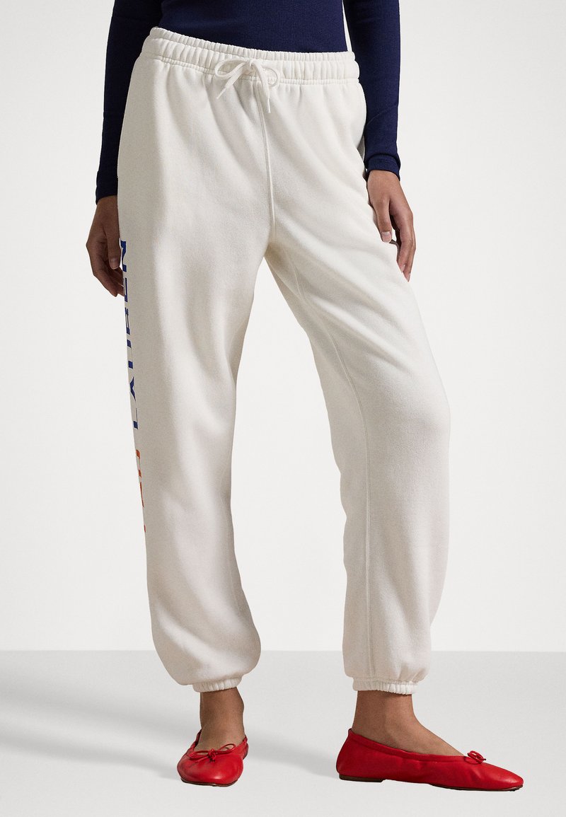 Λευκά παντελόνια φόρμας με ελαστικές μανσέτες και ζώνη, διακοσμημένα με μπλε και πορτοκαλί κείμενο στο πλάι. Συνδυάζονται με κόκκινα μπαλαρίνες.