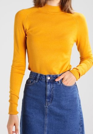 Kobieta ubrana w dopasowany, długi sweter w kolorze musztardowej żółci, wpuszczony w wysokowatą niebieską spódnicę dżinsową z przednimi kieszeniami.