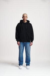 Sudadera negra con cremallera sobre una camiseta negra, combinada con jeans de mezclilla azul claro y zapatillas blancas. El modelo está de pie contra un fondo sencillo.