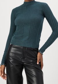 Pull en maille côtelée couleur sarcelle avec col montant et manches longues, orné de boutons décoratifs aux poignets, associé à un pantalon en fausse cuir noir brillant.