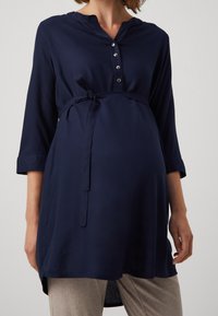 Navy maternity tunic with a buttoned placket, three-quarter sleeves, and a tie belt at the waist. Soft fabric with a smooth texture.