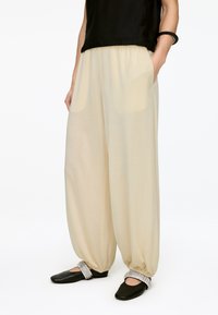 Cremfarbene, weite Hosen aus leichtem Stoff mit einem elastischen Bund und seitlichen Taschen, kombiniert mit schwarzen Ballerinas, die mit Strasssteinen verziert sind.