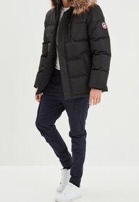 Giacca a vento nera con cappuccio foderato in pelliccia, chiusure a bottoni a pressione e una toppa con logo sul braccio, abbinata a pantaloni cargo blu navy e sneakers bianche.