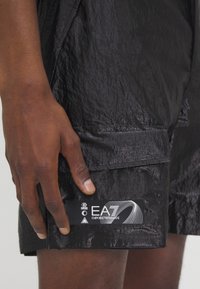 Črne kratke hlače z gubasto teksturo in žepom na obliž. Imajo oznako z logotipom "EA7 Emporio Armani."