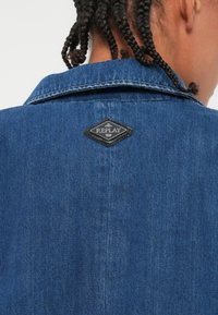 Denim jakna v srednje modri barvi s klasičnim ovratnikom. Ima črn rombeni žep z napisom "REPLAY" na zgornjem delu hrbta.