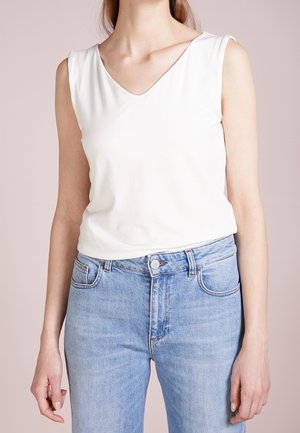 Femme portant un débardeur blanc à col en V rentré dans un jean bleu clair taille haute sur un fond neutre.