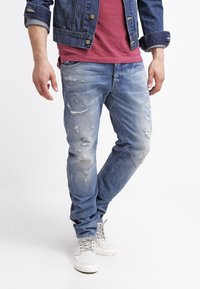 Hombre con jeans claros desgastados, zapatillas blancas, una camiseta roja y una chaqueta de mezclilla oscura, de pie con una pierna cruzada.