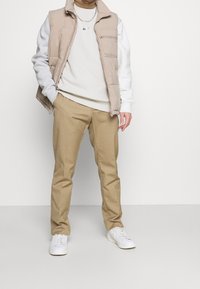 Béžová prošívaná vesta přes bílou mikinu, doplněná světle hnědými kalhotami a bílými teniskami. Výrazným detailem je kapsa na zip.