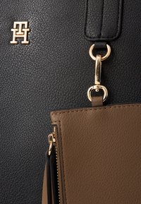 Sort tekstureret lædertaske med en brun accentlomme, der har guldhardware og et logo. Tasken har lynlås-lukning og en aftagelig skulderrem.