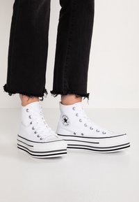 Baskets montantes blanches avec tige en toile, semelle plateforme rayée noire et blanche, et patch logo circulaire sur le côté. Jean noir délavé.