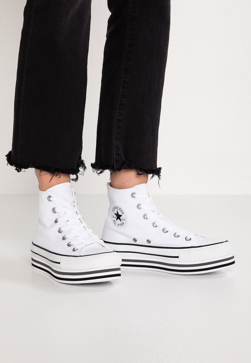 Baskets montantes blanches avec tige en toile, semelle plateforme rayée noire et blanche, et patch logo circulaire sur le côté. Jean noir délavé.