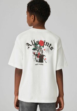 Vit oversized t-shirt med en grafisk design på baksidan som visar en röd ros, ett skelett och en trollkarls hatt, tillsammans med svart text och stjärnor.