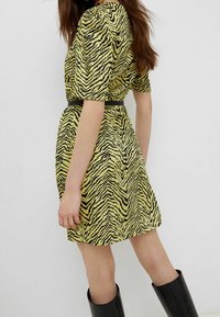 Gelbes Kleid mit einem schwarzen Tigerstreifenmuster. Kurze Ärmel, taillierter Bund und ein fließender Rock. Getragen mit knielangen schwarzen Stiefeln.