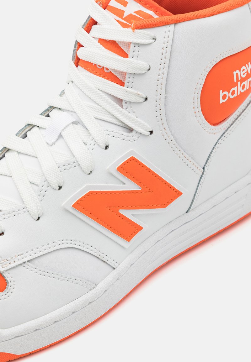 Zapatilla alta blanca con acentos naranjas, que cuenta con una parte superior de cuero texturizado, un logo 'N' prominente y un sistema de cordones reforzado.