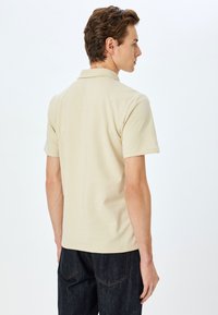 Beige polo skjorte med korte �ærmer, der har en klassisk krave og sideslids. Stof er glat med en let tætsiddende silhuet.