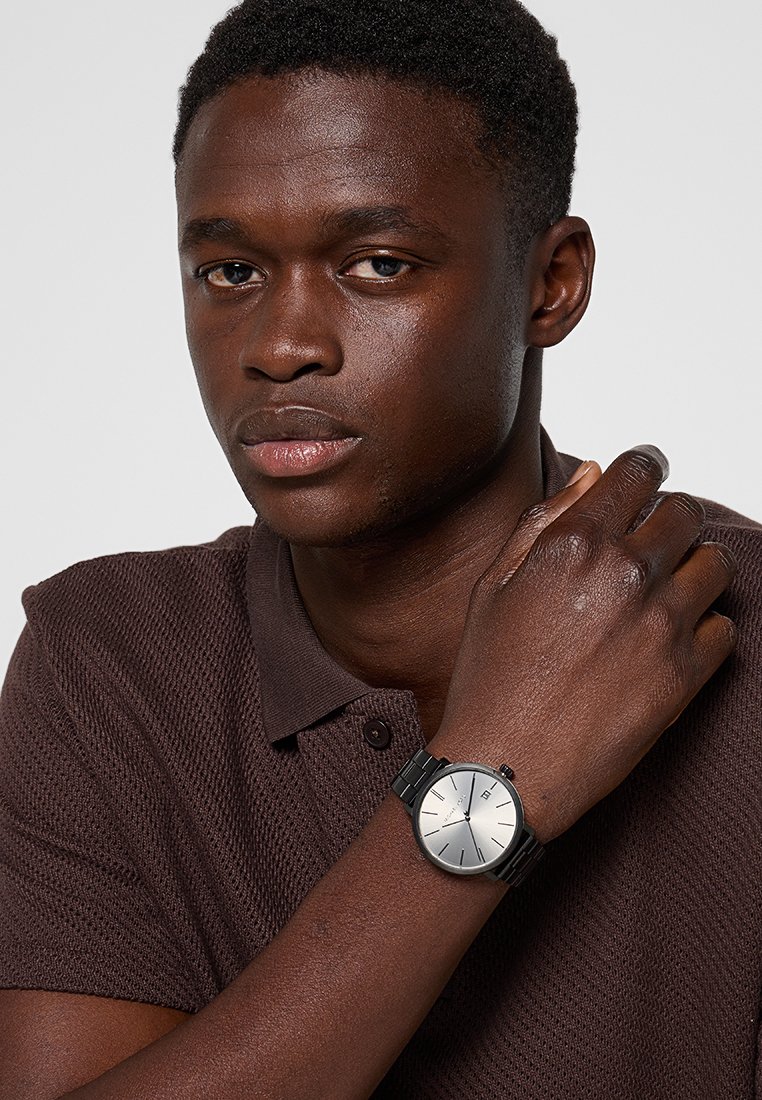 Montre noire avec un cadran argenté, bracelet métallique et design minimaliste. Portée au poignet, associée à un polo texturé marron.