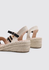 Sandalias de cuña con un upper tejido en beige, correas negras y suela de textura de cuerda. Cuenta con un hebilla ajustable para un mejor ajuste.