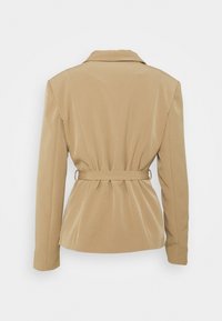 Veste beige cintrée avec manches longues, un col et une ceinture cintrant la taille à l’arrière sur un fond uni.