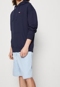 Navy blue hooded sweatshirt in a relaxed fit with a front pocket, featuring a rectangular logo accent on the chest. Worn with light blue shorts.