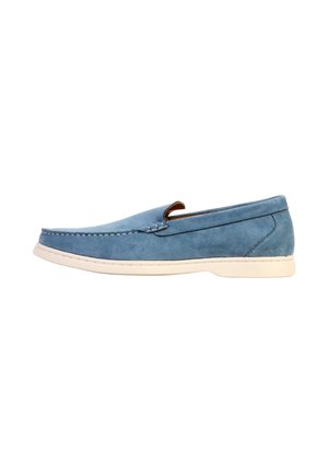 Mocassin bleu en daim à enfiler avec semelle blanche, coutures apparentes le long du bord supérieur et petit talon sur fond blanc.