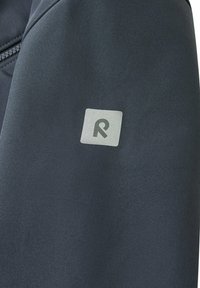 Tissu d'une veste de sport gris foncé avec une texture lisse. Présente un patch logo carré avec une lettre "R" blanche sur la manche.