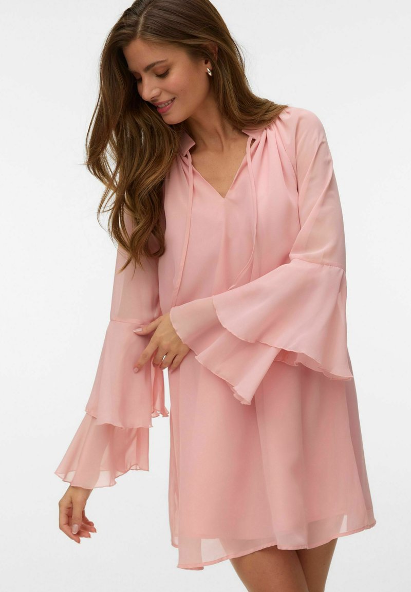 Femme aux longs cheveux bruns portant une robe rose clair fluide avec des manches évasées superposées, souriante et regardant vers le bas.