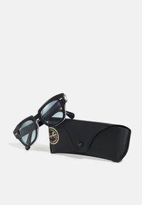 Ochelari de soare Ray-Ban negri, cu rame unghiulare și lentile colorate, așezați pe un toc texturat negru, cu un emblema de logo aurie.