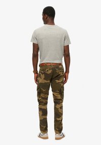 Man standing facing away wearing light gray t-shirt, brown camo cargo pants with belt, white sneakers, and a wristwatch on left wrist.