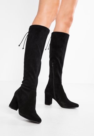 Bottes noires en daim montant jusqu'aux genoux avec talons blocs et fermetures éclair latérales, portées sur des jambes nues sur fond blanc.