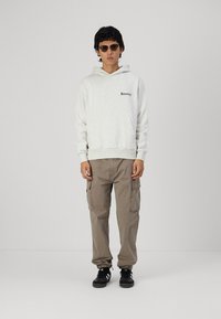Grå hoodie med "Bowery" tekst, lysegrå cargo-bukser og sorte sneakers. Model iført solbriller, stående foran en ensfarvet baggrund.