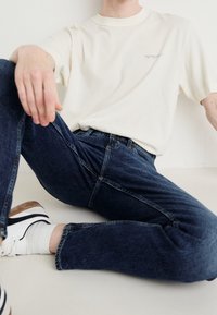 Valkoinen lyhythihainen t-paita yhdistetty tummansiniset farkut. Malli istuu päällään valkoiset sneakerit, joissa on mustia ja ruskeita yksityiskohtia.