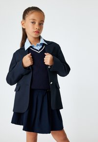 Blazer navy avec boutons argentés, porté sur un gilet navy et une chemise col bleu clair, associé à une jupe navy assortie. Textures lisses.