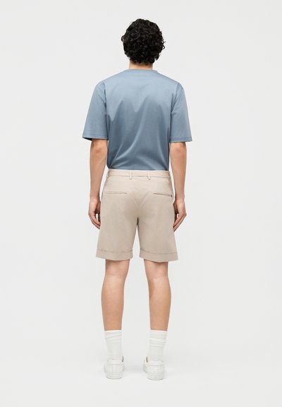 Homme debout de dos, portant une chemise à manches courtes bleue, un short beige, des chaussettes blanches et des baskets blanches sur un fond uni.