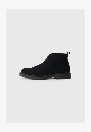Stivali alla caviglia in suede nero con punta rotonda, lacci frontali e una piccola maniglia sul retro. Suola in gomma nera solida.