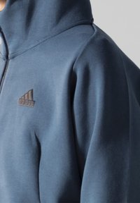 Close-up of a person wearing a blue zip-up hoodie with a small, dark Adidas logo on the chest.