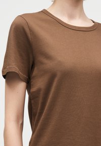 Γυναίκα που φοράει απλό καφέ κοντομάνικο μπλουζάκι με στρογγυλή λαιμόκοψη σε απλό φόντο.